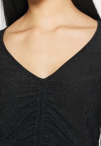 Černý top s výstřihem do V, s nařaseným designem uprostřed a strukturovanou, třpytivou látkou. Žádná viditelná kovová část nebo akcenty.