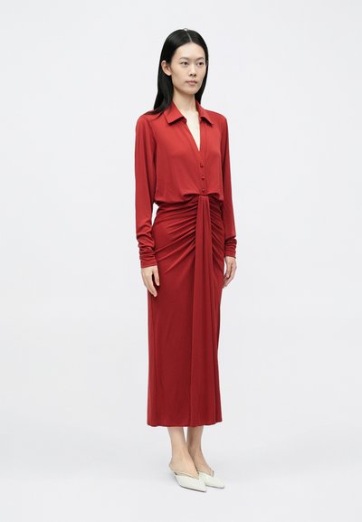 Piros, hosszú ujjú ruha mély V-nyakkivágással, elöl gombolható, deréknál redőzött részletekkel. Sima anyagból készült, bokáig érő fazon.