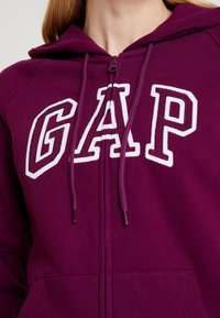 Gros plan sur une personne portant un sweat à capuche zippé maroon de GAP avec des lettres blanches bordées et des cordons, montrant le cou et le haut de la poitrine.
