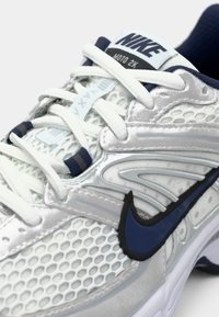 Sneakers Nike Moto 2K mają białą i srebrną siateczkową cholewkę z granatowymi akcentami, teksturowane detale oraz wyraźne czarne logo Swoosh.