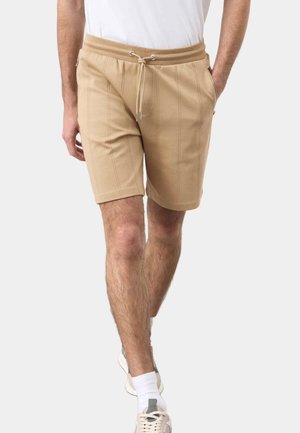 Mann trägt beige knielange Shorts mit Kordelzug und seitlichen Reißverschlusstaschen, weiße Socken und weiße Turnschuhe und geht nach vorne.