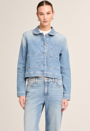 Frau trägt eine hellblaue Jeansjacke mit Fronttaschen und passende hoch taillierte Jeans, steht vor einem beigen Hintergrund.