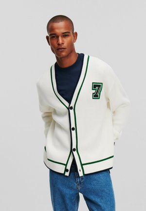 Cárdigan blanco con acentos en verde y marino, diseño de escote en V, botones frontales y un gran parche con el número '7' en el pecho izquierdo. Llevado sobre una camiseta marina.