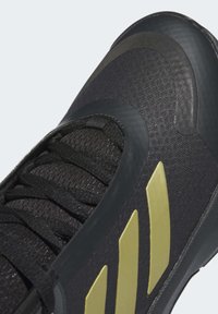 Gros plan d'une basket noire en tissu texturé, lacets noirs et trois bandes diagonales dorées sur le côté.