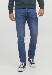 Jeans in denim blu con una vestibilità slim e un leggero effetto delavé, abbinati a scarpe da ginnastica scure con lacci bianchi e suole in gomma.