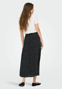 Schwarzer Maxi-Rock mit weißem Blumenmuster, aus leichtem Stoff. Kombiniert mit einem schlichten weißen Oberteil und schwarzen Ballerinas, von hinten gezeigt.