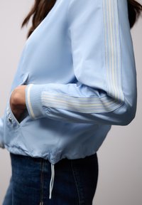 Persona que lleva una chaqueta azul claro con puños y mangas con rayas blancas, con la mano en el bolsillo, combinada con vaqueros de mezclilla azul oscuro.