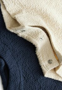 Strickwaren in zwei Farbtönen aus cremefarbenem und nachtblauem strukturiertem Stoff, mit Druckknöpfen auf der cremefarbenen Seite und einem wellenförmigen Muster im ganzen Kleidungsstück.