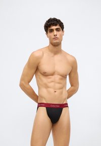 Jonge man met kort, krullend haar, gekleed in zwarte onderbroek met een rode Calvin Klein tailleband, staand tegen een witte achtergrond.