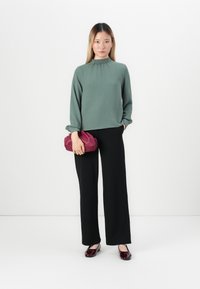 Kvinne som står iført en salviegrønn langermet topp, svarte bukser, burgunderballerinas og holder en matchende burgunder clutchtaske.