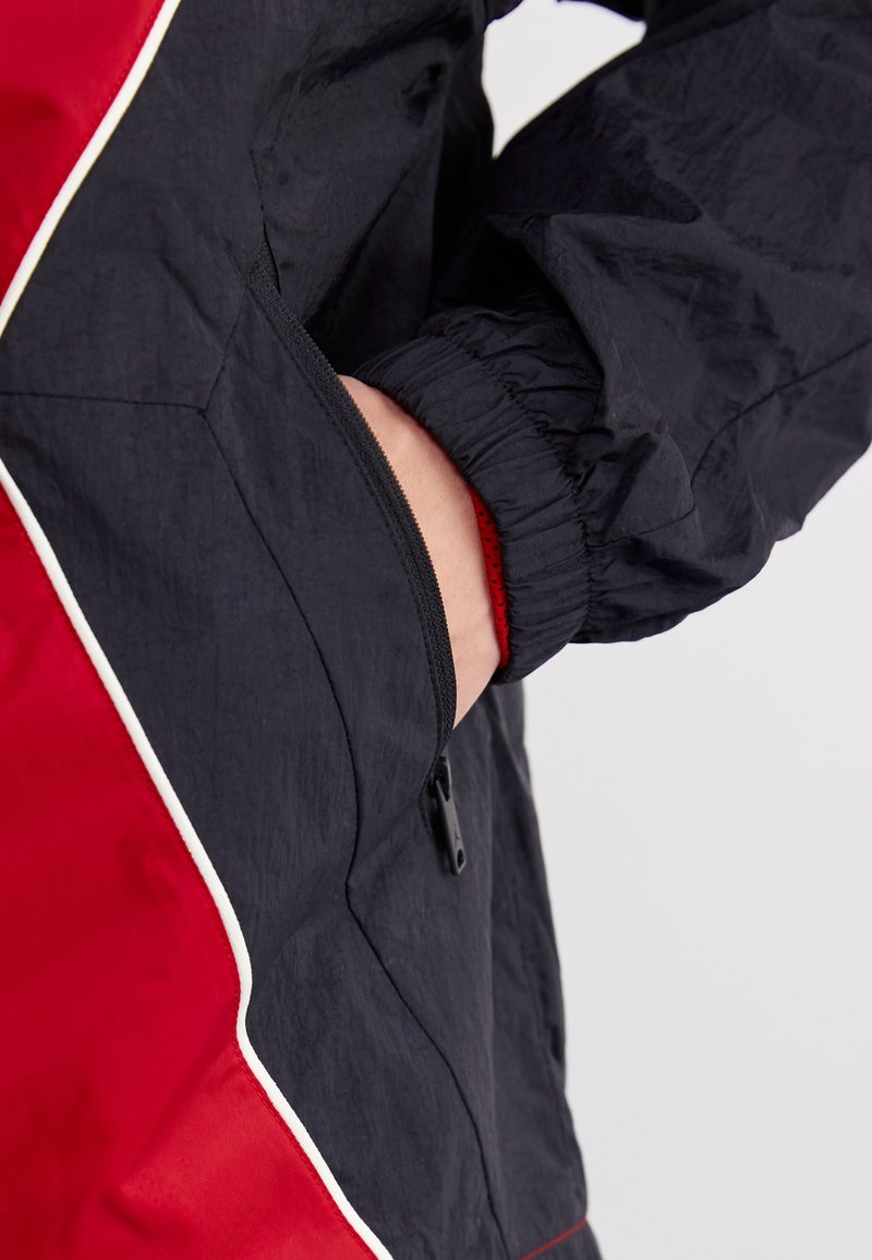 Giacca a vento nera e rossa con tasca laterale, polsini elasticizzati e piping bianco a contrasto lungo le cuciture. Tessuto testurizzato visibile.