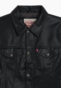 Giacca di pelle nera con design classico, dotata di colletto a punta, tasche frontali, bottoni a pressione e un'etichetta logo rossa.