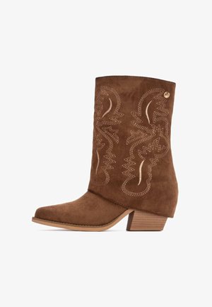 Botín de ante marrón hasta el tobillo con punta afilada, que presenta patrones de costura decorativa y un pequeño tacón cuadrado.