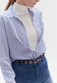 Camicia a righe blu e bianche con colletto aperto sopra un dolcevita bianco, abbinata a jeans a vita alta e una cintura a catena.