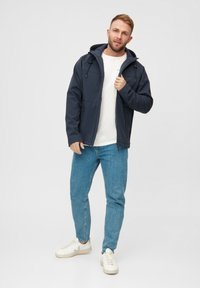 Giacca soft-shell blu marina con cappuccio, zip frontale, due tasche laterali, abbinata a una camicia bianca e jeans azzurri chiari. Sneakers bianche.