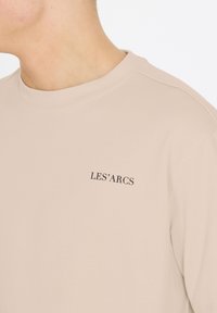 Beige t-shirt med rund halsudskæring og korte ærmer, med sort tekst "LES'ARCS" på brystet. Fremstillet af blødt, glat stof.