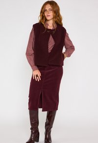 Un gilet borgogna strutturato sopra una camicia a quadri borgogna e bianca, abbinato a una gonna in velluto a coste borgogna con un'apertura frontale e stivali fino al ginocchio.