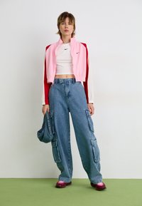 Nike rožinė ir raudona zip-up striukė, balta trumpa palaidinė, mėlyni plačių kojų cargo džinsai ir bordo spalvos batai, laikant mėlyną rankinę.