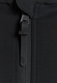 Gros plan d'une fermeture éclair noire avec tissu texturé et tirette marquée du logo "4F" sur un vêtement noir.