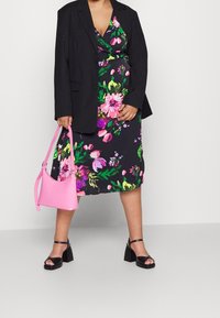 Květinové zavinovací šaty na černém pozadí, zdobené růžovými, zelenými a fialovými vzory. Kombinované s černým blejzrem, růžovou kabelkou a černými sandály.