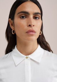 Biely košeľový tričko s hladkou textúrou, s uzatvorením na zlaté gombíky a klasickým dizajnom. Modelka nosí zlaté kruhové náušnice.