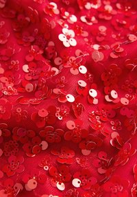Red fabric adorned with numerous small, shiny sequins in floral shapes, creating a textured surface with varied shades of red and reflective accents.