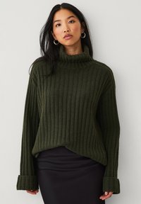 Donkergroene geribbelde turtleneck trui met lange mouwen en brede manchetten. Zachte textuur met een ontspannen pasvorm en een gebogen zoom.