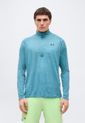 Mees, kes kannab sinist poollukuga spordisärki ja helerohelisi lühikesi pükse, seisab ühevärvilise valge tausta ees.