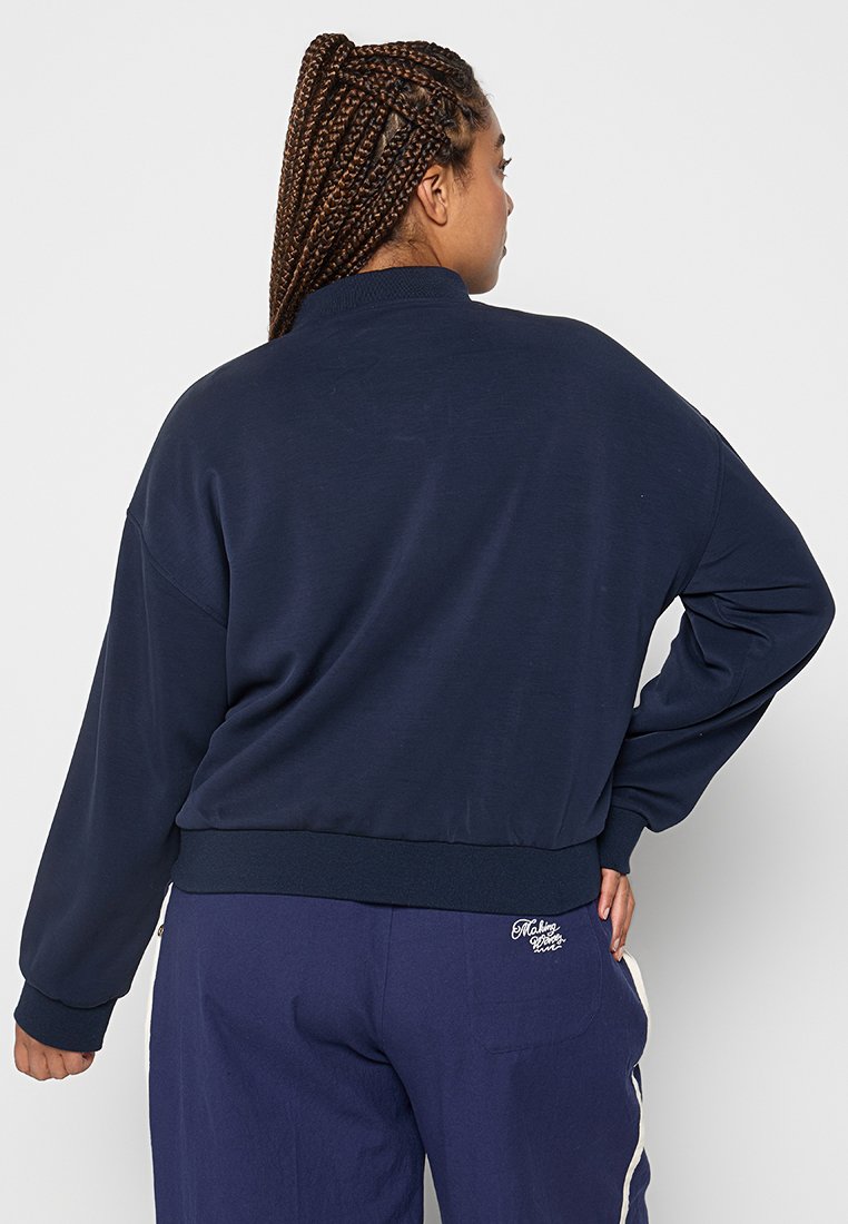 Personne avec de longues tresses portant un sweat-shirt bleu marine et un pantalon assorti avec des rayures blanches sur les côtés, de dos avec une main sur la hanche.
