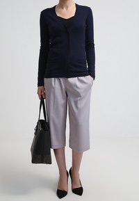 Cardigan blu navy con scollo a V, chiusura a bottoni e due tasche frontali, abbinato a pantaloni grigio chiaro a gamba corta e una borsa tote nera con dettagli borchiati.