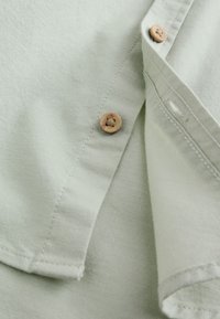 Close-up of light gray fabric with two beige buttons and detailed stitching on a folded shirt sleeve and front placket.