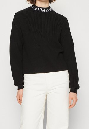 Persona con un suéter negro de punto con el texto "Calvin Klein Jeans" en el cuello y pantalones blancos, de pie contra un fondo liso.