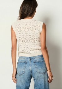Béžový bezrukávý pletený top se vzorem diamantů, žebrovaným výstřihem a lemem, ve spojení s světle modrými džínami s vysokým pasem.