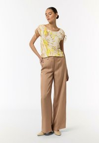 Lässiges Oberteil mit gelbem, weißem und beigem abstrakten Muster. Kombiniert mit beigen, hochgeschnittenen Hosen und beigen Ballerinas. Weicher Stoff.