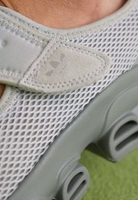 Scarpa sportiva grigia chiara con tomaia in rete, dettagli testurizzati e suola in gomma con fori di ventilazione prominenti. Logo sul lato.