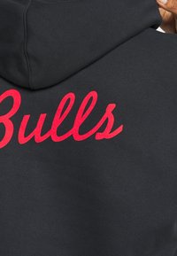 Sweat à capuche noir avec une texture douce, arborant le mot « Bulls » en lettres cursives rouge vif et en gras à l'arrière. Capuche visible en haut.