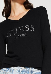 Zwarte V-hals trui van zachte gebreide stof, met een rhinestone 'GUESS' logo en 'EST. 1981' detail op de voorkant. Contrast met lichte denim.