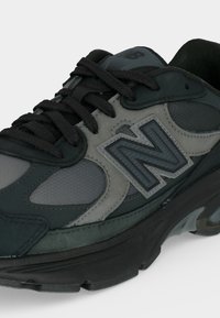 Μαύρο και γκρι αθλητικό παπούτσι New Balance με μαύρα κορδόνια, υφασμένους πλευρικούς πίνακες και παχιά σόλα, φωτογραφημένο με λευκό φόντο.