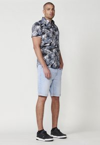 Maglietta nera a maniche corte con motivo tropicale di foglie bianche e azzurro chiaro, abbinata a pantaloni corti di jeans azzurri e scarpe da ginnastica nere.