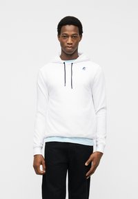 Νεαρός άνδρας που φοράει λευκό φούτερ με μαύρα κορδόνια, ανοιχτό γαλάζιο πουκάμισο από κάτω και μαύρο παντελόνι, στέκεται μπροστά σε ένα απλό ανοιχτό φόντο.