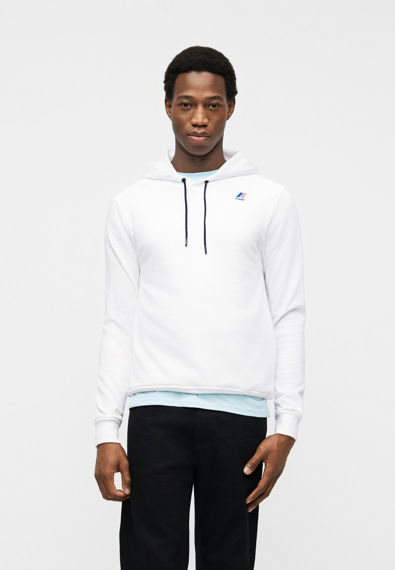 Νεαρός άνδρας που φοράει λευκό φούτερ με μαύρα κορδόνια, ανοιχτό γαλάζιο πουκάμισο από κάτω και μαύρο παντελόνι, στέκεται μπροστά σε ένα απλό ανοιχτό φόντο.