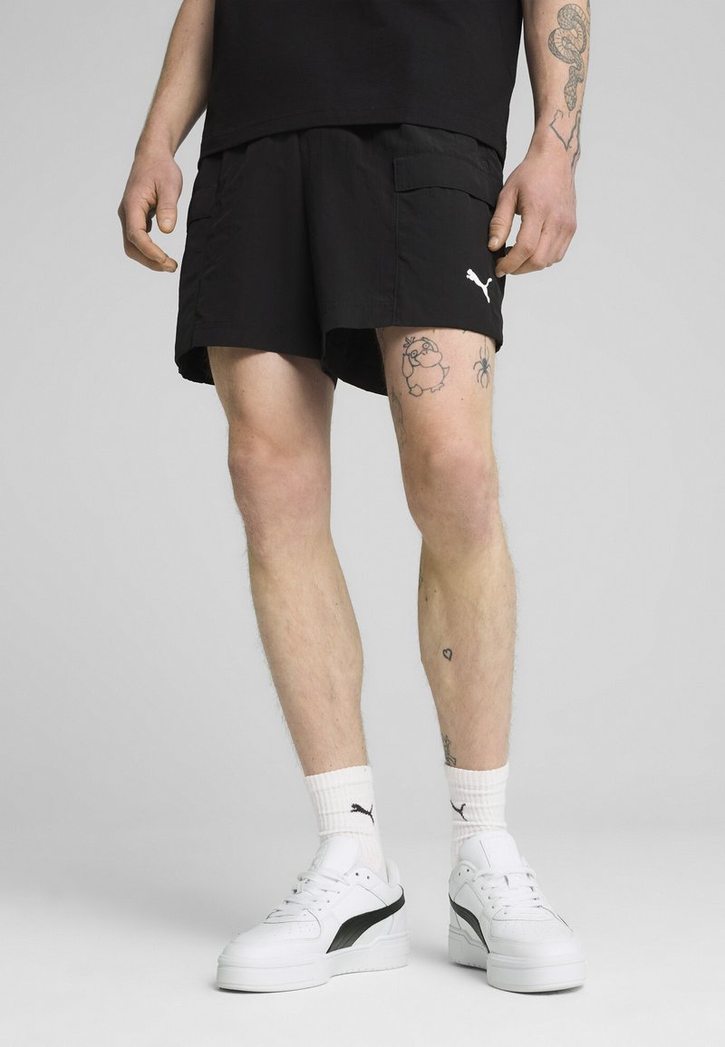 Shorts de sport noirs avec poches latérales et un logo Puma blanc, associés à des baskets blanches avec des accents noirs et des chaussettes côtelées.