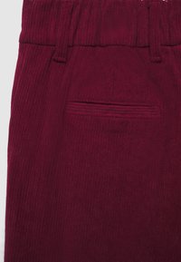 Nahaufnahme eines burgunderfarbenen Cordstoffkleidungsstücks mit Gürtelschlaufen und einer horizontalen Einschubtasche.