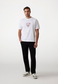 Hvid bomulds t-shirt med grafisk print, der viser "Guess" og "Los Angeles, Californien." Sammen med sorte bukser og sneakers.