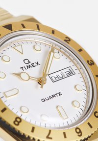 Orologio al quarzo dorato con quadrante bianco, grandi indicatori delle ore, lancette dorate e display della data. Presenta una lunetta girevole e una finitura liscia.