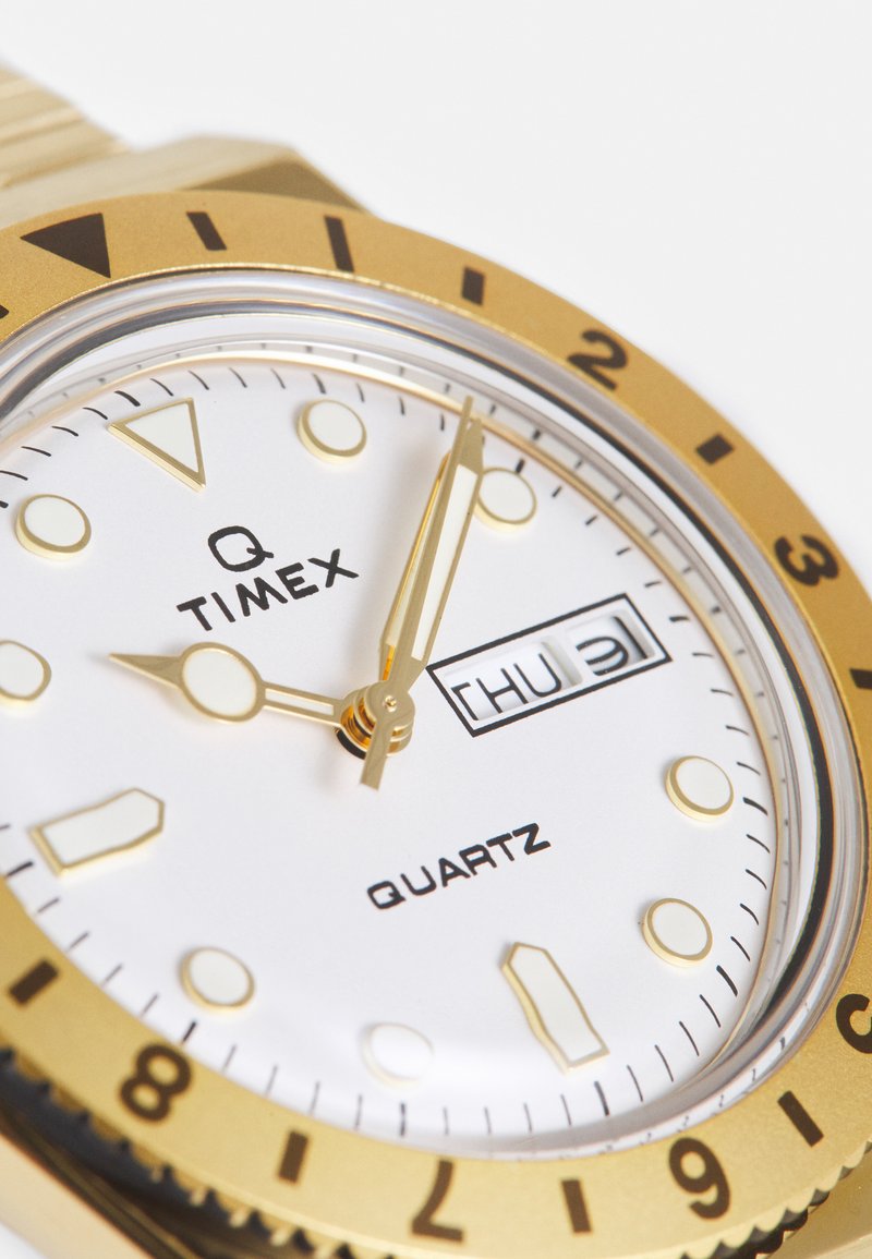 Reloj de cuarzo dorado con un dial blanco, grandes marcadores de horas, manecillas doradas y una pantalla de fecha. Presenta un bisel giratorio y un acabado suave.