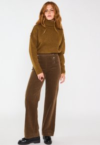 Pantaloni a gamba larga in corduroy marrone con dettagli in bottone, abbinati a un maglione a collo alto testurizzato color senape. Modella in piedi su uno sfondo neutro.