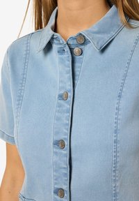 Camisa de denim azul claro com mangas curtas, apresentando um colarinho e cinco botões metálicos na parte da frente. Textura suave.
