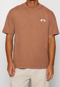 Mann trägt ein braunes Kurzarm-T-Shirt mit kleiner weißer Elch-Stickerei auf der linken Brust, kombiniert mit hellbeigen Hosen.