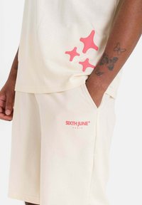 Beige cotton t-shirt with pink star-shaped accents, paired with matching shorts featuring a logo in pink. Soft texture, casual design.